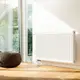 Weißer Buderus-Heizkörper mit gerillter Oberfläche an einer hellen Wand neben einem großen Fenster, das Tageslicht hereinlässt. Ein runder Hocker steht auf dem Holzboden.