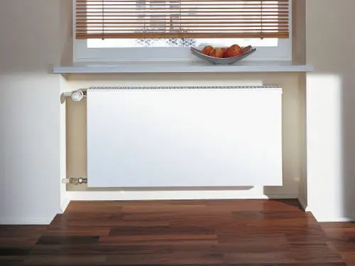 Weißer Kermi-Heizkörper, flach und horizontal, montiert unter einem Fenster. Die Oberfläche ist glatt und befindet sich in einem modernen Raum mit Holzfußboden.