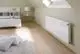 Ein weißer Heizkörper vor einer beigen Wand in einem Schlafzimmer mit Bett und Teppich.