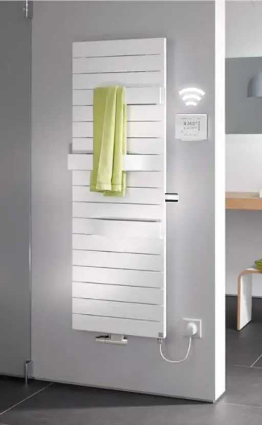Vertikaler Kermi-Heizkörper in Weiß mit horizontalen Rippen, montiert an einer hellen Wand, mit einem Handtuch und einem angrenzenden Steuergerät sichtbar.