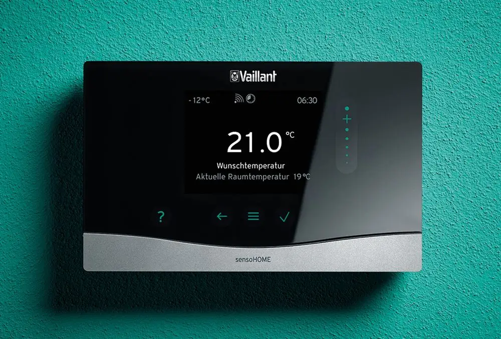 Raumtemperaturregler von Vaillant mit digitalem Display, Anzeige der Wunsch- und aktuellen Temperatur, Steuerungselemente und eBUS-Schnittstelle, montiert an einer grünen Wand.