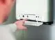 Daikin-Wärmepumpe an einer Wand montiert, mit einem Bedienfeld, das Tasten und Anzeigen zeigt. Eine Hand drückt auf einen Knopf am Gerät.