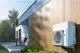 Daikin Klimaanlage, wandmontiert, mit Außengerät. Das Gerät ist an einer Holzverkleidung montiert, im Hintergrund ist ein Garten mit Rasen und Bäumen sichtbar.