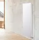 Vertikaler Heizkörper von HM Heizkörper in weißer Farbe, montiert an einer betongrauen Wand, sichtbar in einem modernen Raum mit Holzfußboden und Fensterblick nach draußen.