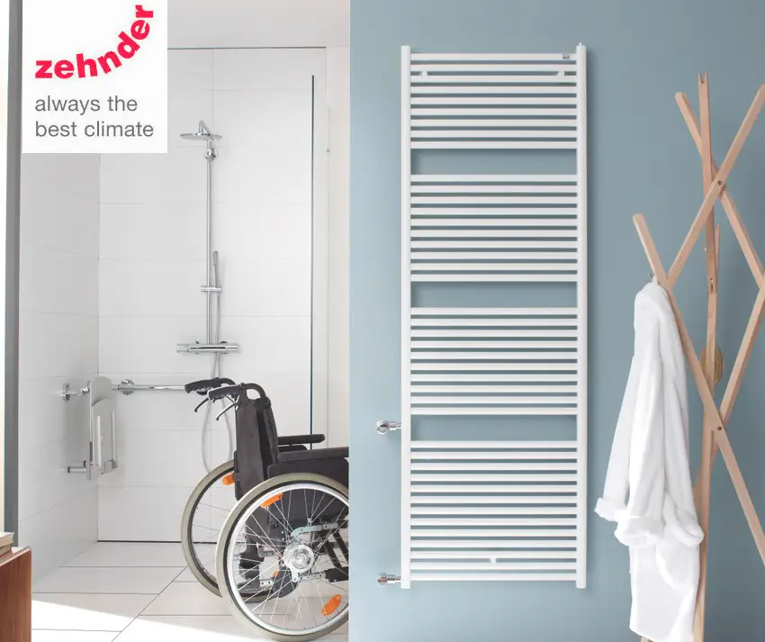Zehnder-Heizkörper in vertikaler Ausführung, weiß mit Längsrippen, montiert an einer Wand in einem modernen Badezimmer mit Dusche und Rollstuhl.