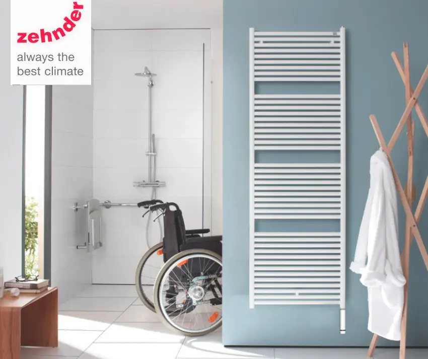 Weißer Heizkörper von Zehnder mit Längsrippen an einer blauen Wand im Badezimmer. Im Hintergrund ist eine Dusche und ein Rollstuhl zu sehen.