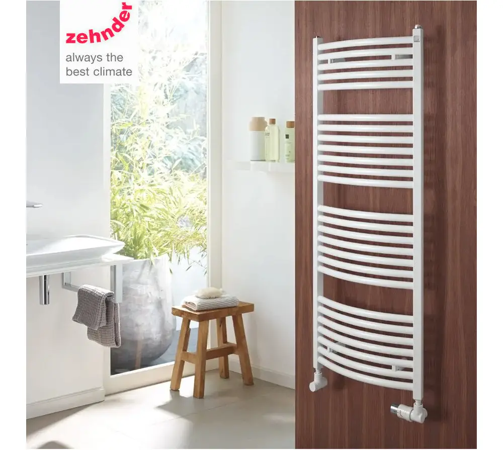 Weißer Badheizkörper von Zehnder mit gebogener Oberfläche, montiert an einer Holzwand, neben einem Waschbecken und einem Hocker. Badezimmer mit Fenster und Pflanzen im Hintergrund.