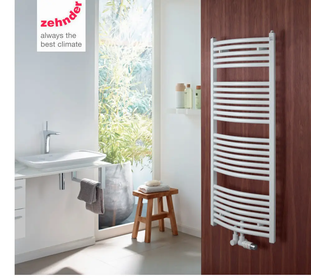 Zehnder Badheizkörper in weißer Farbe, gebogen, an einer Holzwand montiert, neben einem Waschbecken, mit Handtuchhalter und Fensterausschnitt im Hintergrund.