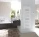 Vertikaler Elektroheizkörper von Zehnder in Weiß, montiert an einer hellen Wand, neben einem modernen Badezimmer mit Badewanne und Waschbecken.