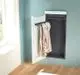 Zehnder Elektroheizkörper in vertikaler Montage, installiert in einem Raum, mit hellblauer Wand und natürlichem Licht von einem Fenster, hält zwei Kleidungsstücke.