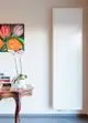 Vertikaler, weißer Heizkörper von Purmo an der Wand, neben einem Tisch mit Pflanzen und bunten Büchern, und einem blühenden Gemälde im Hintergrund.