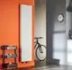 Purmo Heizkörper, vertikal montiert, in Weiß an einer orangefarbenen Wand, neben einem Fahrrad und einem Regal mit Obstschalen.