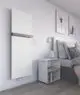 Purmo Heizkörper in vertikaler Position, weiß, an einer hellen Wand montiert. Neben einem Nachttisch mit Bücherregal und einer dekorativen Vase im Schlafzimmer.