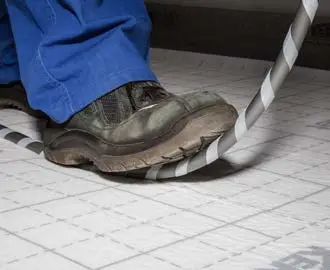 Füße in Arbeitsschuhen stehen auf einem Boden mit einem Kabel, das teilweise um den Schuh gewickelt ist, umgeben von grauen Fliesen. Kermi wird für die Wärme im Raum genutzt.