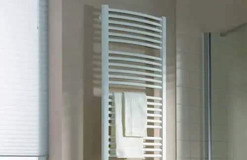 Weißer Handtuchheizkörper mit mehreren Lüftungsstreben und einem aufgehängten Handtuch, vor einer beige gestrichenen Wand und einem transparenten Duschabtrennung.