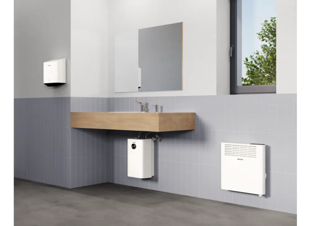Badansicht mit Holzwaschtisch, zwei Wasserhähnen, einem weißen Heizkörper und einer Stiebel Eltron Sensor-Armatur. Wand und Boden in neutralen Farben mit Fensterlicht.