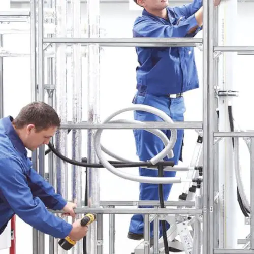 Zwei Handwerker in blauen Overalls installieren Rohrleitungen in einem gerüsteten Rahmen, einer nutzt ein Elektro-Werkzeug, der andere arbeitet auf einer Leiter.