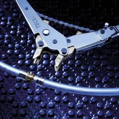 Eine technische Zange mit Kettenmechanismus und einem transparenten Schlauch mit einer goldenen Verbindung auf einer strukturierten, dunklen Oberfläche.