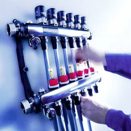 Ein Rohrsystem mit mehreren Ventilen und Reglern ist an der Wand montiert. Eine Hand dreht einen der Regler. Die Komponenten sind glänzend und modern gestaltet.