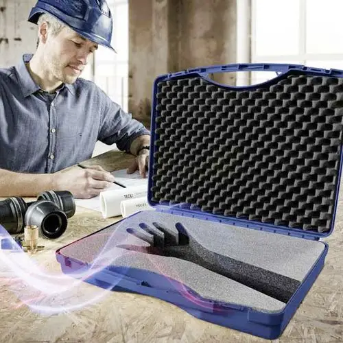 Ein Arbeiter mit Schutzhelm schreibt Notizen, während eine geöffnete blaue Box mit Schaumstoffeinlage und einer Handform daneben liegt. Rohrsystemteile sind im Vordergrund sichtbar.