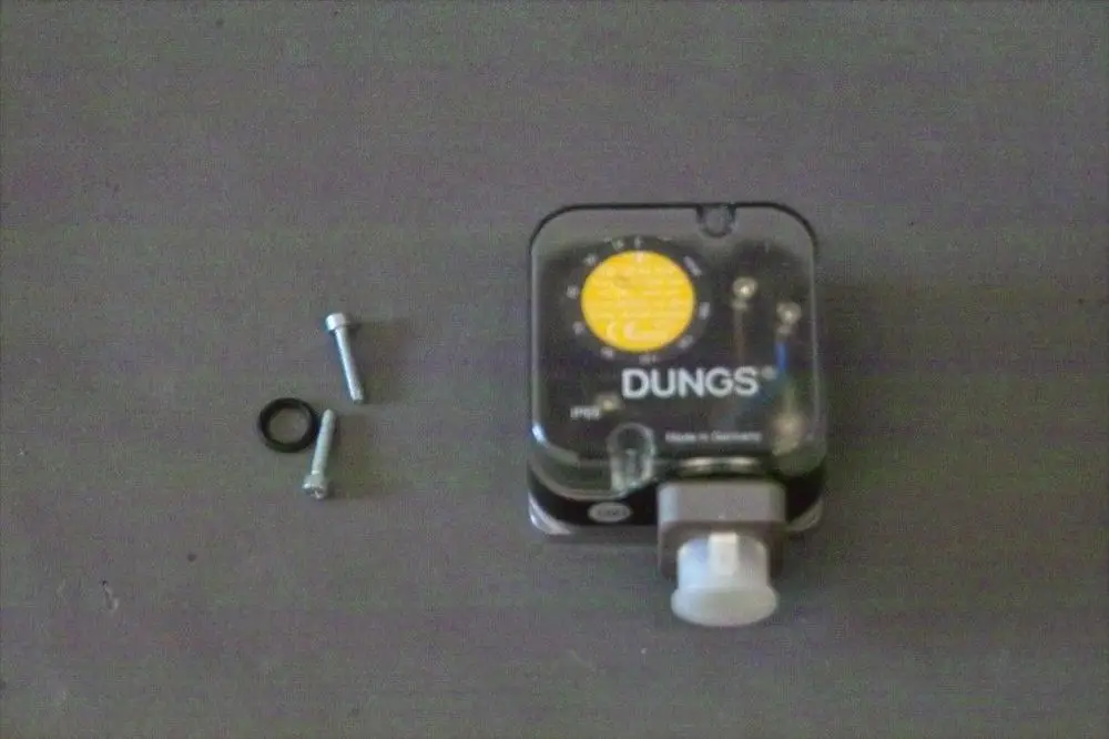 Dungs Gasdruckwächter mit transparentem Gehäuse, Ventil und Anschlussstück, begleitet von zwei Schrauben und einem Gummiring, auf einem grauen Untergrund.