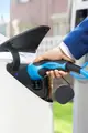 Hand, die ein blaues Mennekes-Ladegerät in eine weiße Elektroauto-Ladebuchse einführt. Im Hintergrund sind Ladesäulen und eine grüne Rasenfläche sichtbar.