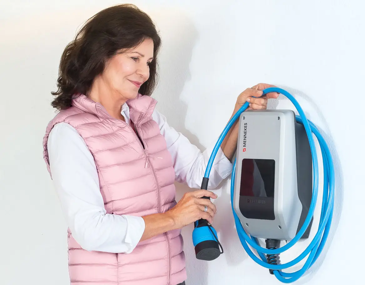 Frau mit dunklen Haaren in einem rosa Steppwesten hält ein Kabel in der Hand. An der Wand ist eine Mennekes-Ladestation für Elektrofahrzeuge montiert.