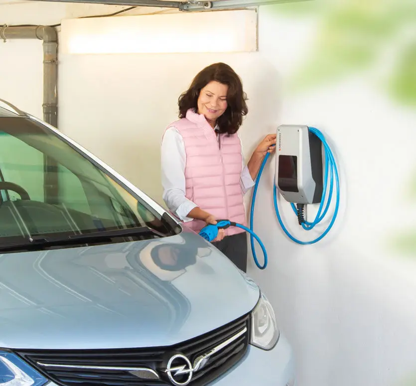 Frau verbindet ein Elektrofahrzeug über ein blaues Ladekabel mit einer Wandladestation von MENNEKES in einer Garage.