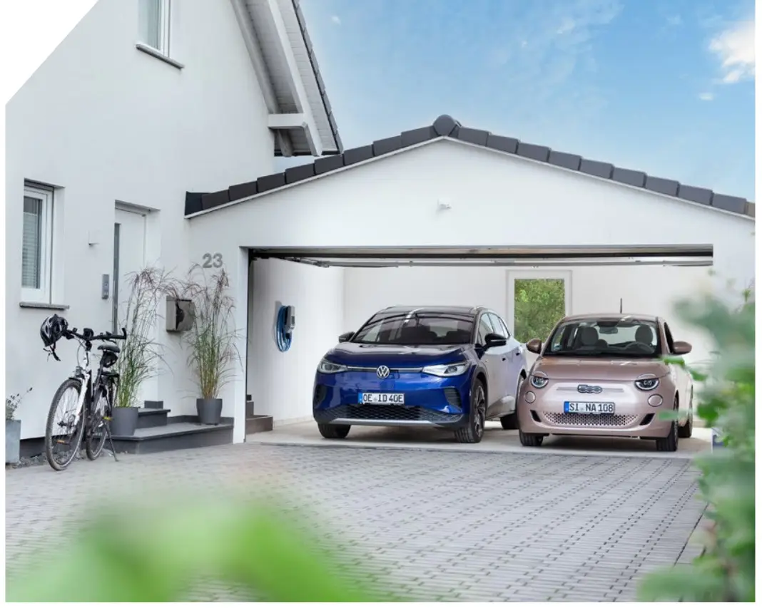 Zwei Elektroautos stehen in einer Garage, neben einem Mennekes-Ladeanschluss an der Wand. Ein Fahrrad ist auf der linken Seite abgestellt. Helle Fassade und Pflastersteine im Vordergrund.
