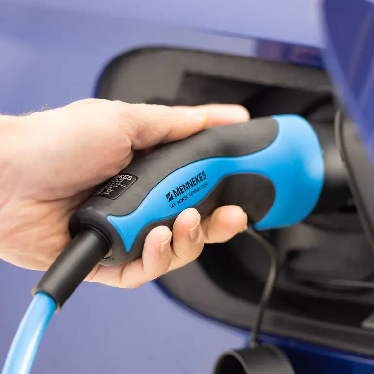 Hand mit einem blauen und schwarzen Mennekes-Stecker, der in eine Elektroladestation eingeführt wird. Der Stecker hat eine ergonomische Form und einen elektrischen Kabelanschluss.