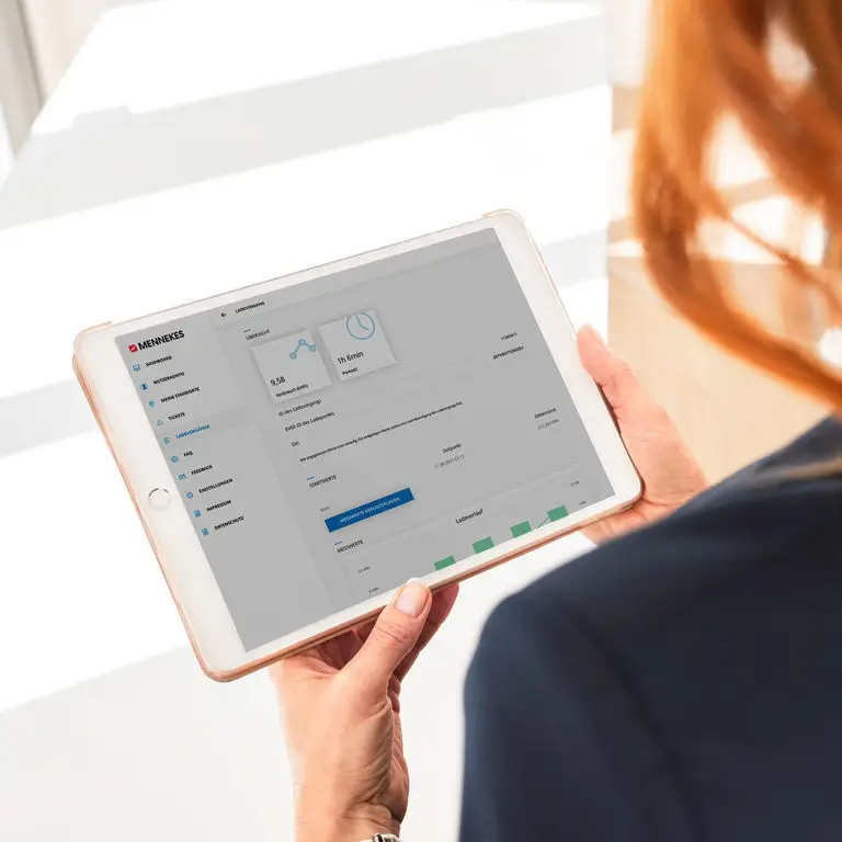 Tablet in Händen einer Person, auf dem die Mennekes Softwareoberfläche zu sehen ist, einschließlich Diagrammen und Leistungsdaten. Heller Hintergrund und professionelles Setting.