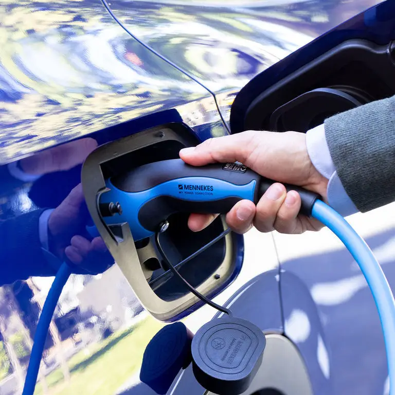 Hand steuert einen MENNEKES-Ladeanschluss, der mit einem Elektrokabel verbunden ist, während eine blaue Fahrzeugkarosserie im Hintergrund sichtbar ist.