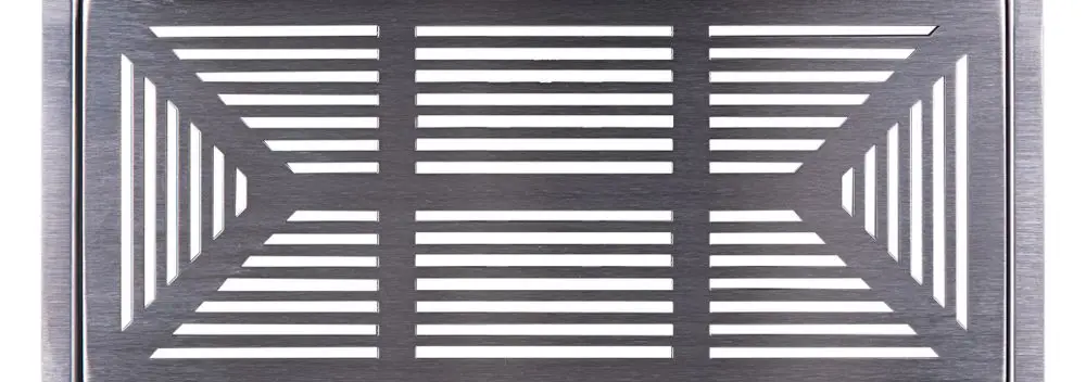 Edelstahl-Abdeckgitter mit einem geometrischen Streifenmuster, platziert auf einem flachen Untergrund. Das Produkt stammt von der Marke Pluggit.