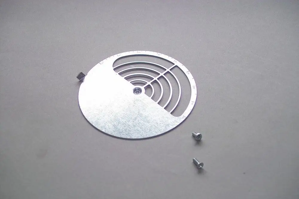 Metallische Luftklappe von Brötje mit kreisförmigem Design, Ausschnitten und Skalenmarkierungen. Zwei Schrauben liegen daneben auf einer grauen Oberfläche.
