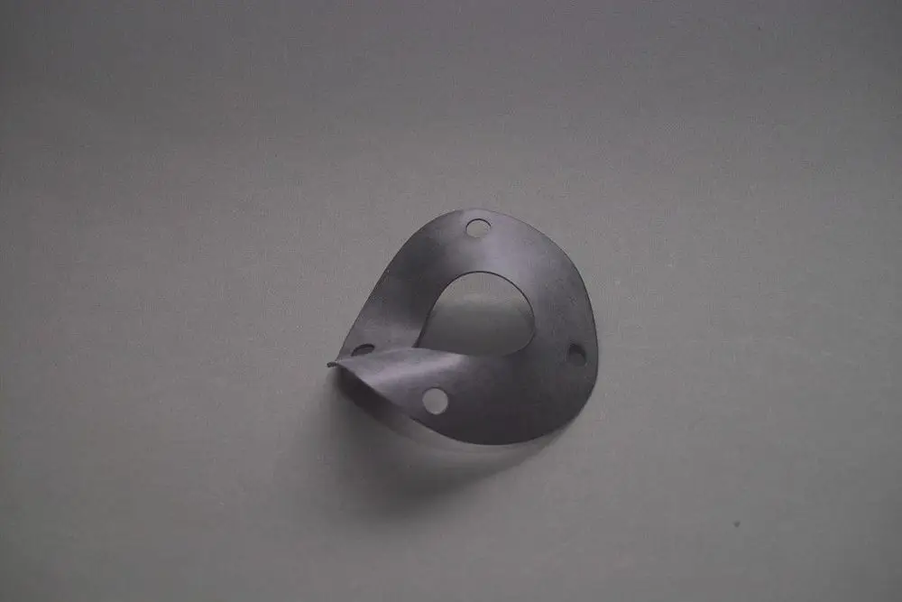 Brötje Metalldichtung, geformtes Metallprofil mit einer Öffnung in der Mitte und drei Bohrungen, auf grauem Hintergrund platziert.