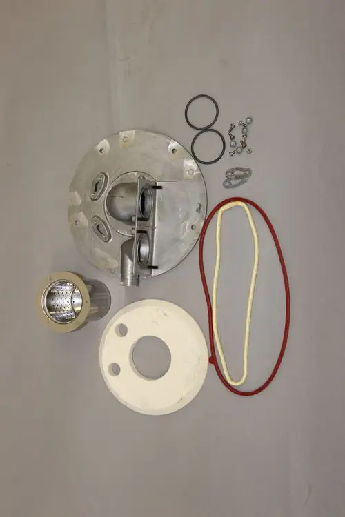 Brötje Brennerdeckel mit mehreren Bauteilen, darunter O-Ringe, Schrauben und Filter, auf grauem Untergrund angeordnet.