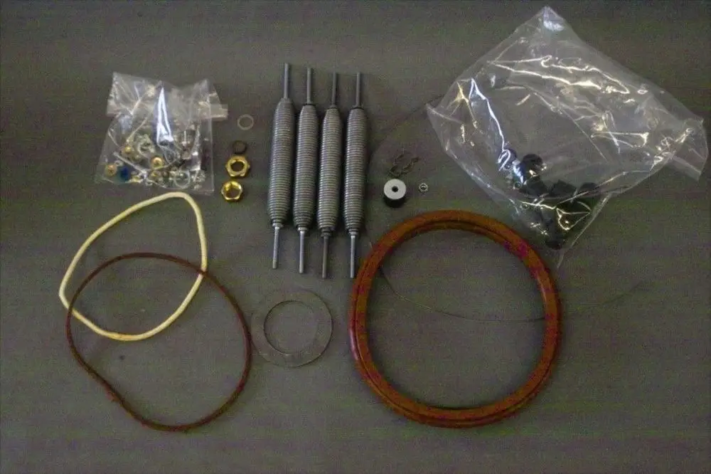 Brötje Feder-Set mit mehreren Federn, Gummibändern, Schrauben, Unterlegscheiben und weiteren Kleinteilen in verschiedenen Verpackungen auf grauem Untergrund.