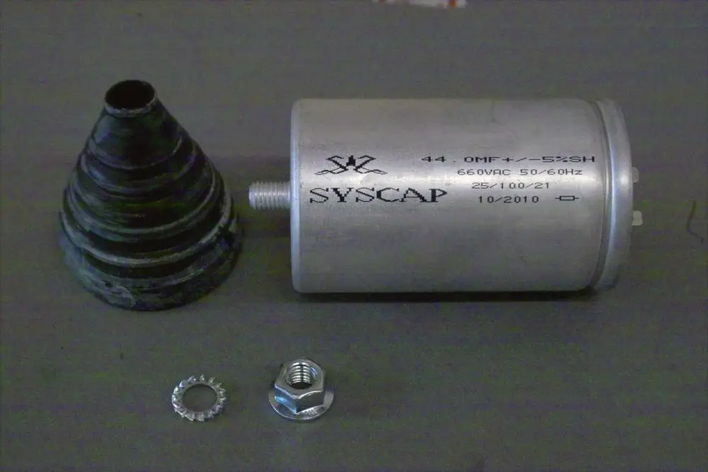 Kondensator von Brötje in zylindrischer Form, mit Metallgehäuse und Anschlussgewinde, neben einem konischen Gummi-Element und Metallverschlüssen auf grauem Hintergrund.