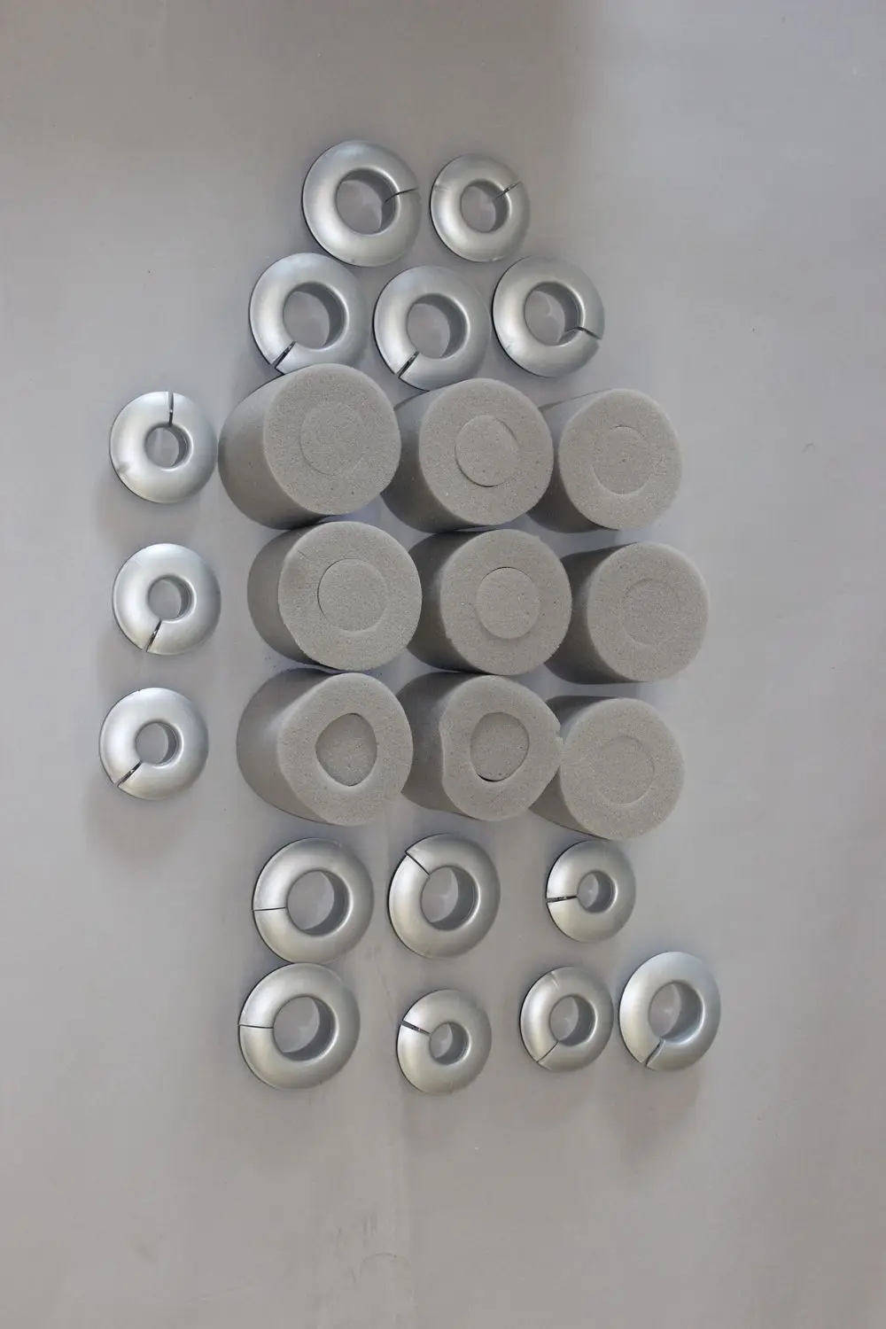 Anordnung von grauen Schaumstoffzylindern und silbernen, ringförmigen Metallteilen auf heller Fläche, möglicherweise Zubehörteile von Brötje.