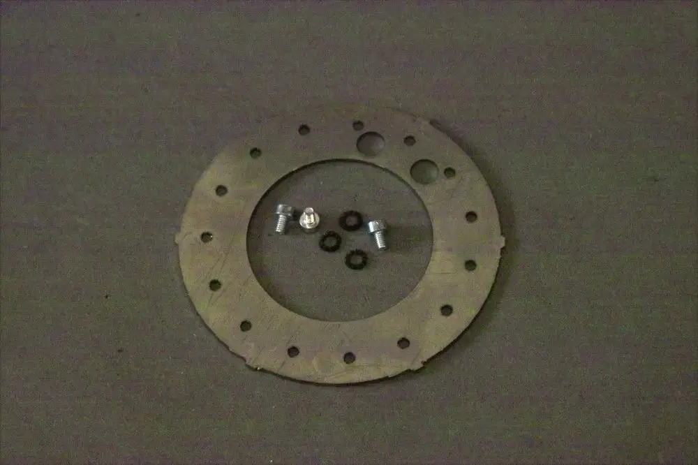 Metallische Stauscheibe mit mehreren Löchern, darauf liegend zwei Schrauben und vier Dichtungen, auf einem grauen Untergrund. Produkt von Brötje.