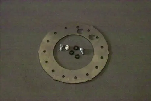 Metallische Stauscheibe mit mehreren Löchern, darauf liegend zwei Schrauben und vier Dichtungen, auf einem grauen Untergrund. Produkt von Brötje.