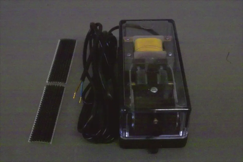 Brötje Booster, ein elektrisches Gerät mit transparentem Gehäuse und zwei Anschlusskabeln. Daneben befinden sich zwei kleine, gezahnte Elemente.