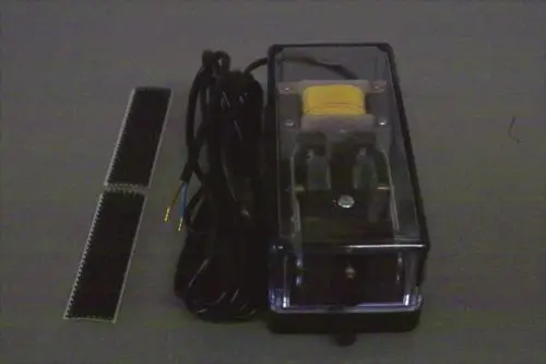 Brötje Booster, ein elektrisches Gerät mit transparentem Gehäuse und zwei Anschlusskabeln. Daneben befinden sich zwei kleine, gezahnte Elemente.