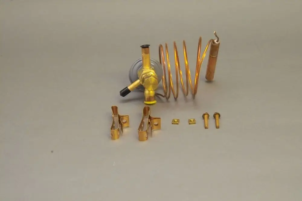 Brötje Expansionsventil mit einer Kupferschlange, einem Kugelhahn und mehreren Montageschrauben sowie Clips im Bild angeordnet auf grauem Untergrund.