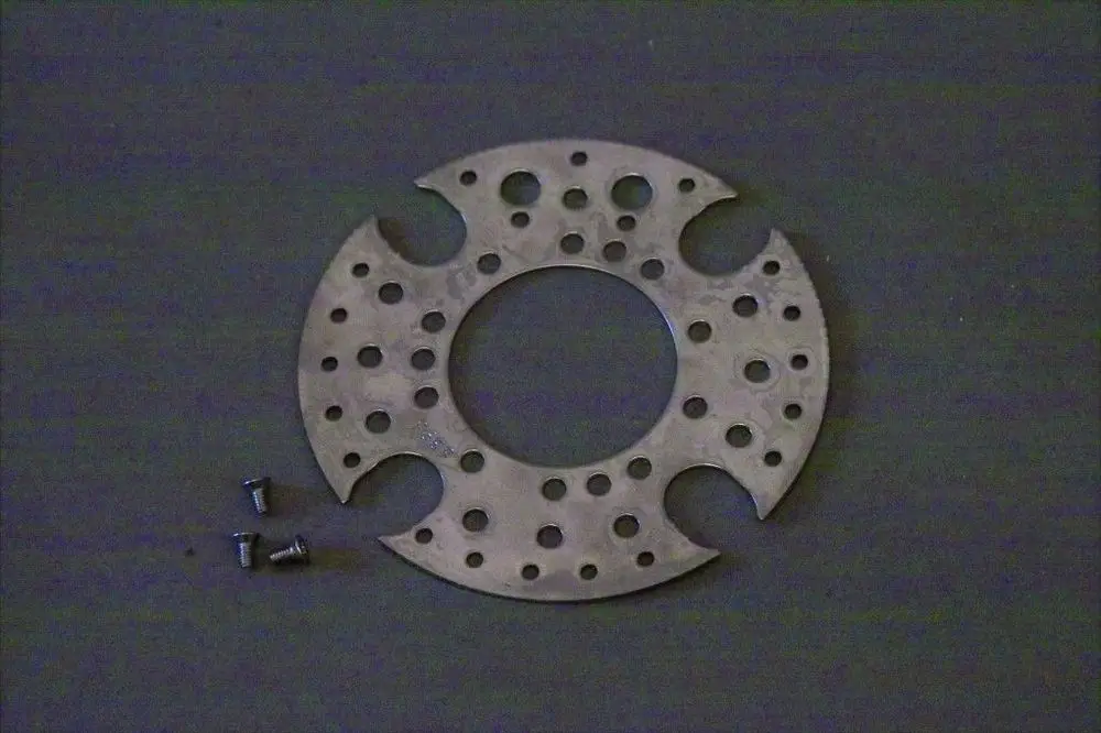 Metallene Stauscheibe mit mehreren Löchern und Aussparungen, liegend auf einer dunklen Oberfläche. Dazu mehrere kleine Schrauben. Hersteller ist Brötje.