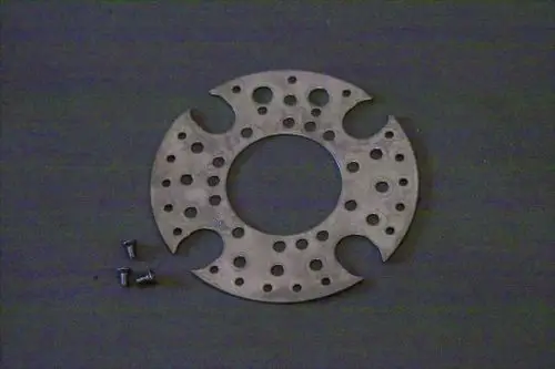 Metallene Stauscheibe mit mehreren Löchern und Aussparungen, liegend auf einer dunklen Oberfläche. Dazu mehrere kleine Schrauben. Hersteller ist Brötje.