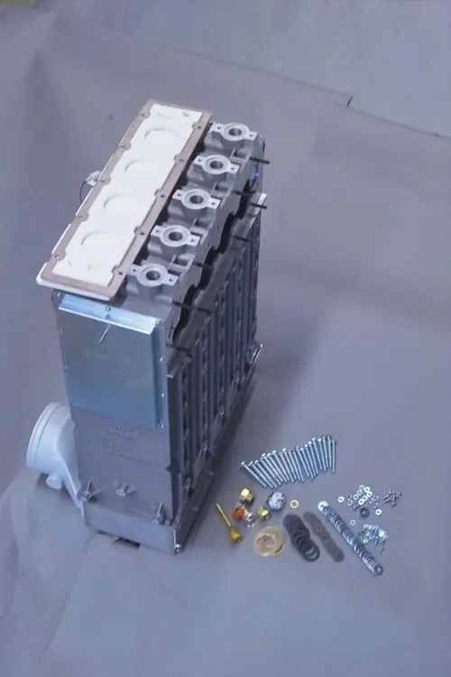 Brötje Wärmetauscher mit Anschlussseelen und Zubehörteilen auf grauem Untergrund, bestehend aus Schrauben, Dichtungen und weiteren Bauteilen.