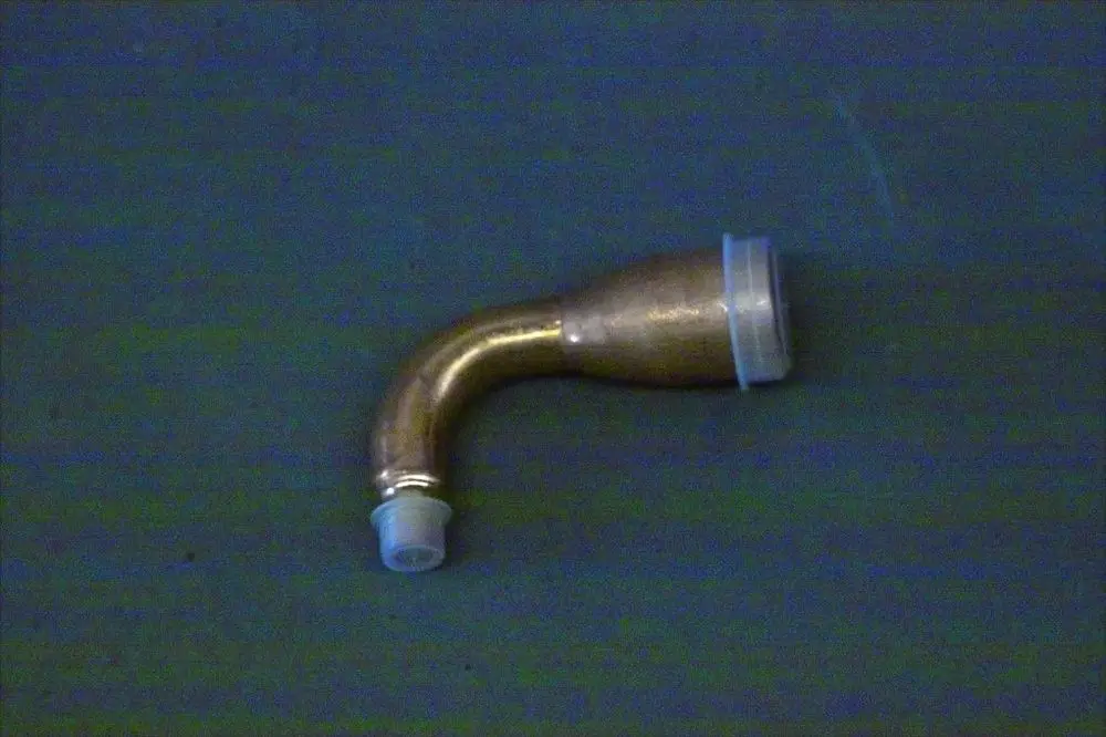 Eingeknickte Einspritzleitung aus Messing mit transparentem Endstück, auf einer unifarbenen Unterlage. Produkt von Brötje zur Verwendung in Heizsystemen.