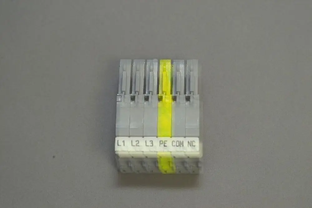 Brötje Stecker mit sechs Anschlüssen, beschriftet mit L1, L2, L3, PE, COM und NC. Das Produkt hat eine graue Farbe und eine senkrechte Ausrichtung.