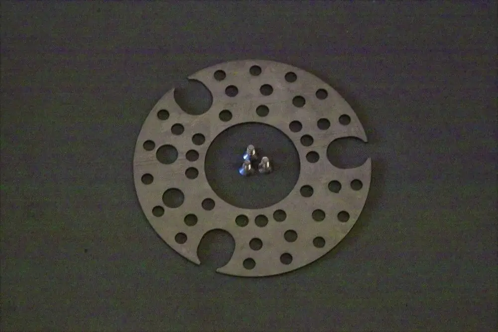 Brötje Stauscheibe mit mehreren Löchern und Einschnitten, in der Mitte angeordnet, umgeben von drei kleinen Metallkugeln auf dunklem Hintergrund.